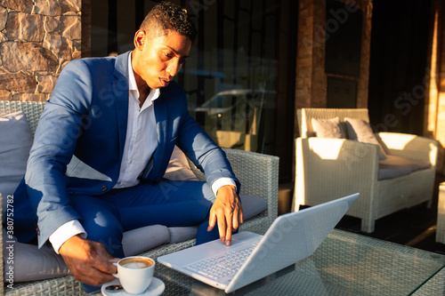 Young African American male entrepreneur in formal wear working on laptop and thinking about financial problem while sitting in lounge zone of modern cafeteria photo