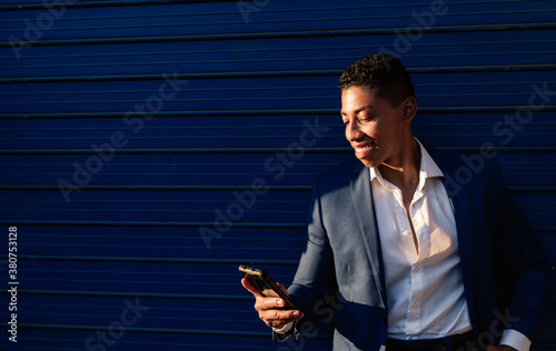 Happy young African American male manager in elegant formal clothes reading message on smartphone and smiling  while standing against dark blue wall on street photo