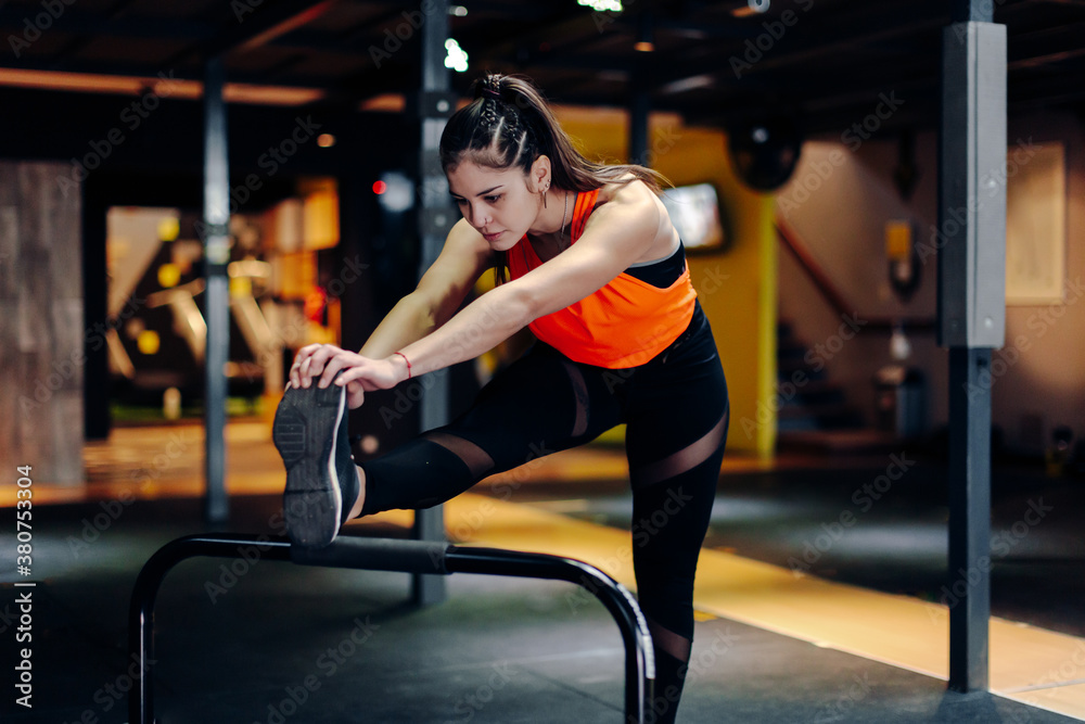 Full body young sporty female in fitness outfit performing stretching exercise near metal bar while warming up during training in gym Full body young sporty female in fitness outfit performing stretching exercise near metal bar while warming up during training in gym