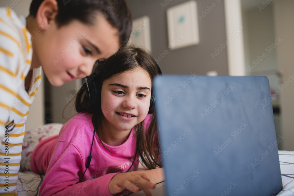 Happy little sister and brother having fun and playing game on laptop together while spending free time in bedroom at home Happy little sister and brother having fun and playing game on laptop together while spending free time in bedroom at home