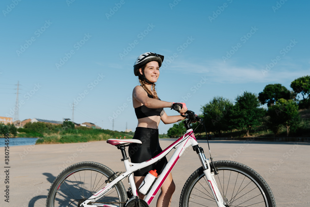 Side view of positive young female cyclist in black sportswear and protective helmet standing with bike on paved road and looking away confidently Side view of positive young female cyclist in black sportswear and protective helmet standing with bike on paved road and looking away confidently
