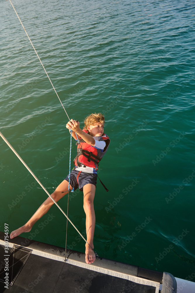 Vertical photo of a blond boy with life jacket holding a rope with his feet on the edge of a boat sailing in the middle of the sea Vertical photo of a blond boy with life jacket holding a rope with his feet on the edge of a boat sailing in the middle of the sea