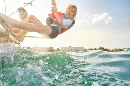 Blond boy with a life jacket laughing as he grabs a rope from a boat to make counterweights hanging out to sea with a man next to him sitting on the boat steering the wheel