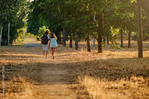 Two kids with school bags and a mask on their backs walking far away along a path in the woods while talking