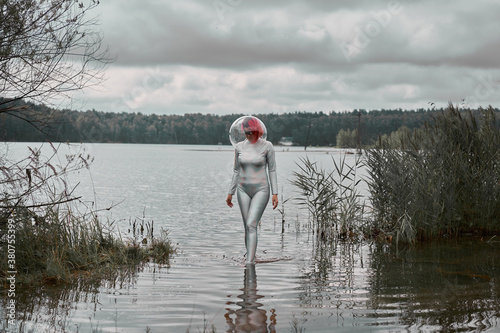 Futuristic young red haired female in silver space suit and glass helmet walking out of flooding river photo