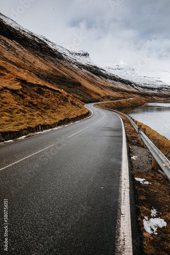 Spectacular view of wet winding road located near river and mountain slope with dry grass in winter on Faroe Islands