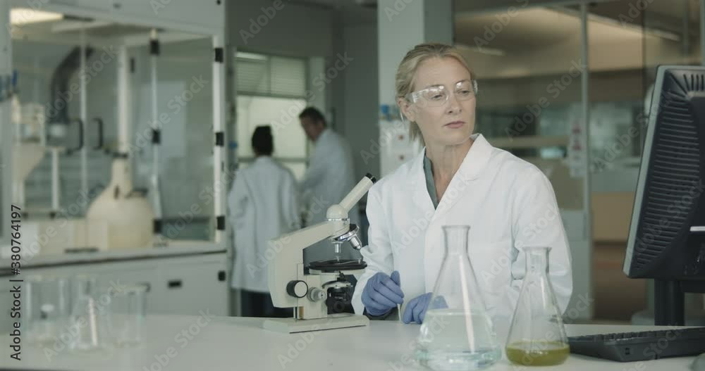 Mature Female Laboratory Scientist Conducts Experiment in Science lab looking at microscope in busy lab at night and working on computer Mature Female Laboratory Scientist Conducts Experiment in Science lab looking at microscope in busy lab at night and working on computer