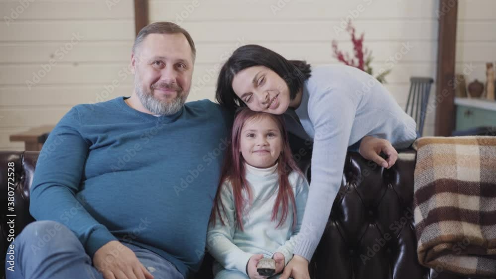 Happy family posing in living room at home. Portrait of smiling Caucasian father, mother and daughter resting on weekends. Cheerful people looking at camera and smiling. Happy family posing in living room at home. Portrait of smiling Caucasian father, mother and daughter resting on weekends. Cheerful people looking at camera and smiling.