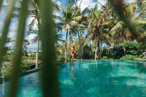 Girl in red swimsuit on the edge of pool in the jungles with ocean on the background. Palms around and crystal clean water. Luxury resort on Bali island