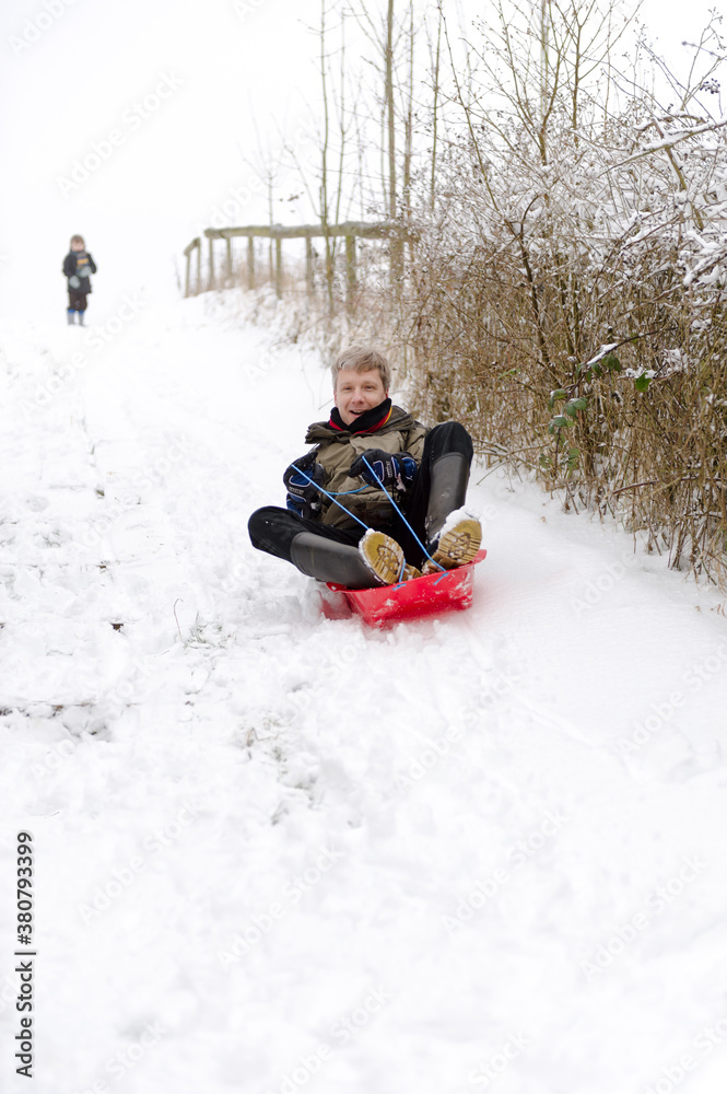 Man in kids sled with son chasing behind Man in kids sled with son chasing behind