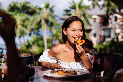 Happy asian pretty woman hungry having pizza at sunny day  sunset light in outdoor restaurant. Female enjoying  food  having fun at lunch. Tropical background.  