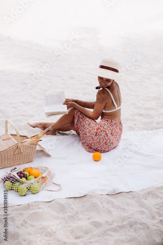Romantic portrait of stylish asian woman sit on beach carpet blanket having picnic with basket of fruits, wearing top, long skirts and big large straw hat, holding reading book. 