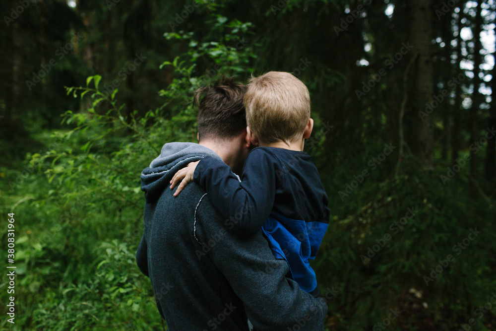 An teenager holds a younger boy (seen from the back) as they examine plants in a forest. An teenager holds a younger boy (seen from the back) as they examine plants in a forest.