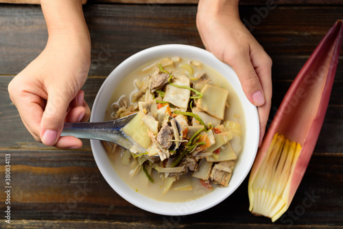 Northern Thai food (Kaeng Hua Plee), banana blossom spicy soup with pork in a bowl holding by hand and eating on wooden table, Local Thai food photo