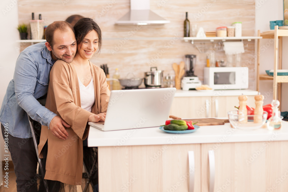 Cheerful couple using laptop in kitchen reading online recipe for breakfast. Happy loving cheerful romantic in love couple at home using modern wifi wireless internet technology Cheerful couple using laptop in kitchen reading online recipe for breakfast. Happy loving cheerful romantic in love couple at home using modern wifi wireless internet technology