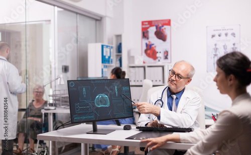 Doctor with grey hair pointing at x-ray on computer for young patient in hospital office. Young woman with severe injury on brain after car accident. Disabled mature woman in wheelchair talking with
