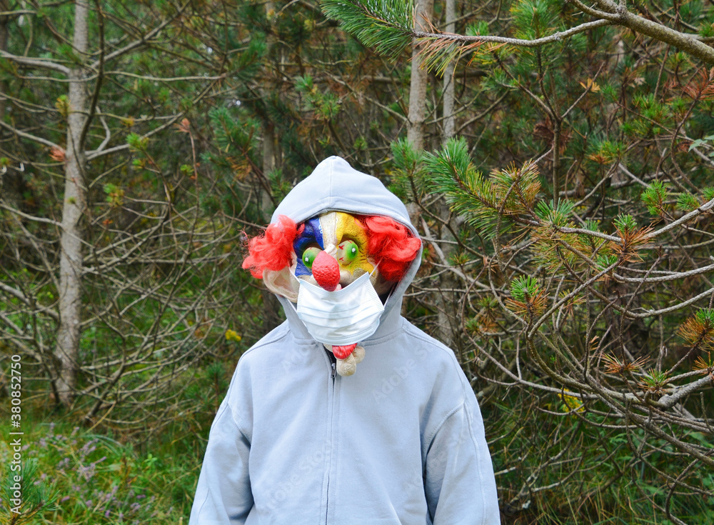 Boy on his back with a hooded jacket in the forest who, when he turns around, wears a mask from the movie It and in his mouth a mask from the covid. Halloween party with security measures.