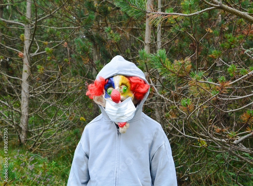 Obraz na płótnie Boy on his back with a hooded jacket in the forest who, when he turns around, wears a mask from the movie It and in his mouth a mask from the covid. Halloween party with security measures. w salonie
