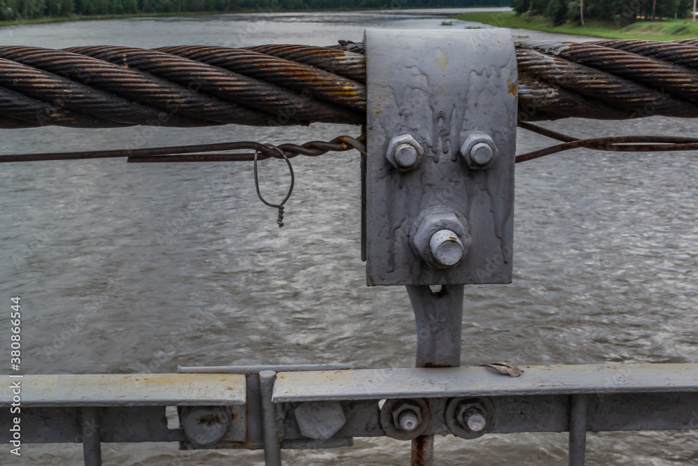 Gray metal shackle connect the sling and rope tied knot. Close up details of metal parts of suspension footbridge. River backdrop