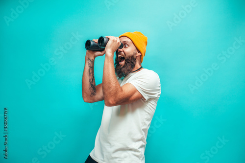 Young bearded hipster guy with binoculars over isolated blue wall wearing white blank t-shirt. Crazy emotions photo
