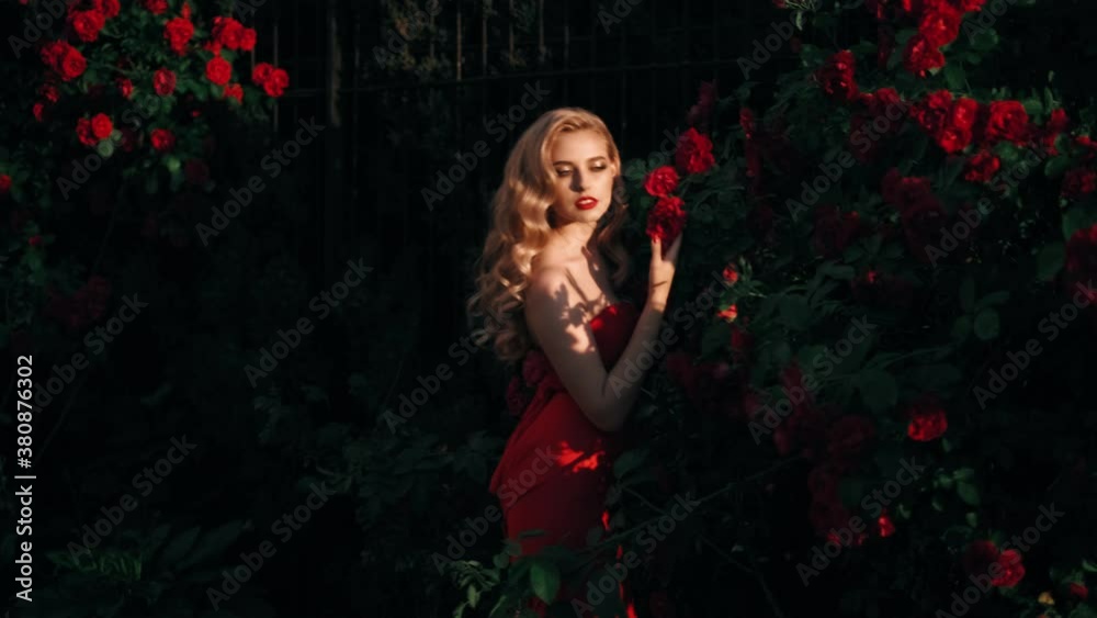 Fabulous girl with red lips in red dress on awesome summer roses background. Fantasy woman portrait. Awesome blonde model expresses emotions. Luxury lady in red is flirting. Beauty and style concept Fabulous girl with red lips in red dress on awesome summer roses background. Fantasy woman portrait. Awesome blonde model expresses emotions. Luxury lady in red is flirting. Beauty and style concept