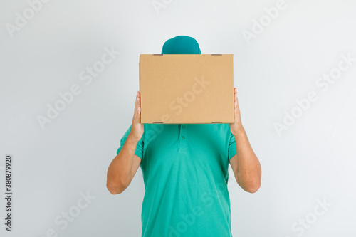 Delivery man in green t-shirt, cap holding cardboard box over face , front view.