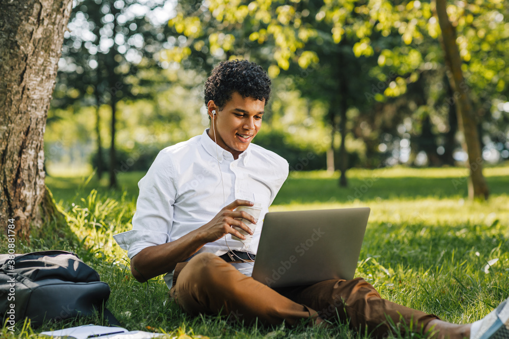 Smiling african american college boy using a laptop computer sitting in park. Young man looking on laptop and drinking coffee. Smiling african american college boy using a laptop computer sitting in park. Young man looking on laptop and drinking coffee.