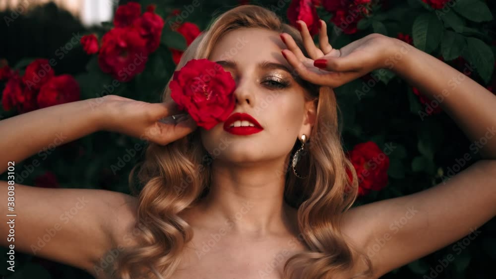 Fabulous girl with red lips in red dress on awesome summer roses background. Fantasy woman portrait. Awesome blonde model expresses emotions. Luxury lady in red is flirting. Beauty and style concept Fabulous girl with red lips in red dress on awesome summer roses background. Fantasy woman portrait. Awesome blonde model expresses emotions. Luxury lady in red is flirting. Beauty and style concept