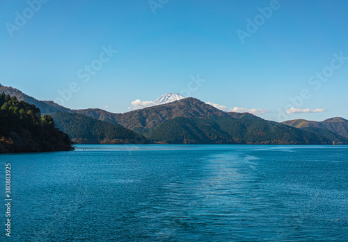 Photo from the boat on the lake in the morning, sunlight shines on the water. There is a blue sky with fluffy white clouds. The Mount Fuji with snow on the peak and trees behind. There is copy space.