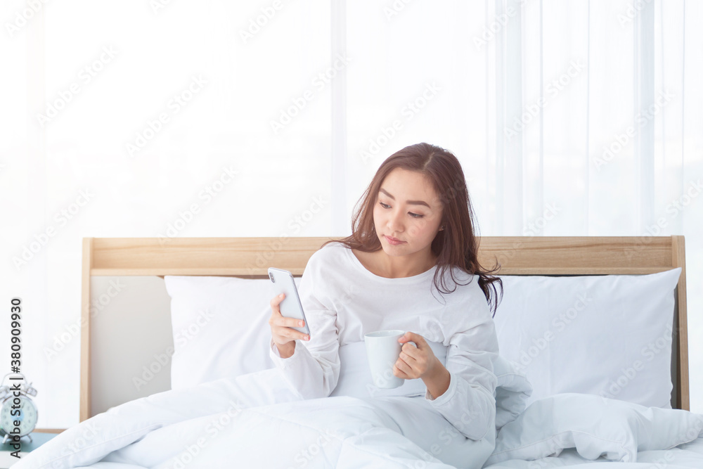 Morning girl with cup of coffee in hands in bed on a white blanket, simple, home, while playing with smartphone. Beautiful Asia woman drinking a coffee in her bed while reading from smartphone. Morning girl with cup of coffee in hands in bed on a white blanket, simple, home, while playing with smartphone. Beautiful Asia woman drinking a coffee in her bed while reading from smartphone.