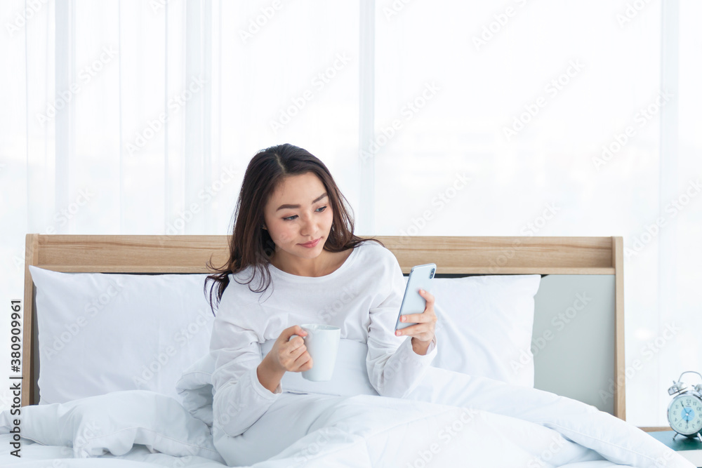 Morning girl with cup of coffee in hands in bed on a white blanket, simple, home, while playing with smartphone. Beautiful Asia woman drinking a coffee in her bed while reading from smartphone. Morning girl with cup of coffee in hands in bed on a white blanket, simple, home, while playing with smartphone. Beautiful Asia woman drinking a coffee in her bed while reading from smartphone.