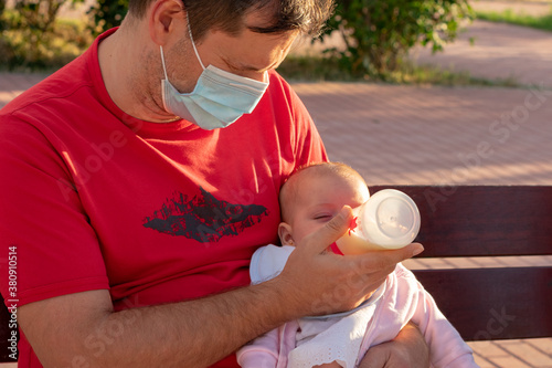 Primer plano de un padre sentado en un banco de un parque. El padre que lleva una mascarilla quirúrgica da el biberón a su bebé.