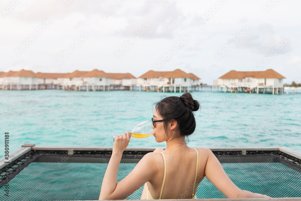Young beautiful girl in a hammock with wine glass over the blue sea. Young girl enjoying glass of wine by the sea. Young woman watching sunset from sea hammock enjoying tropical vacations.