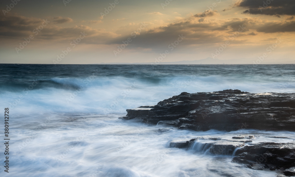 Rocky seashore with wavy ocean and waves crashing on the rocks at sunset.