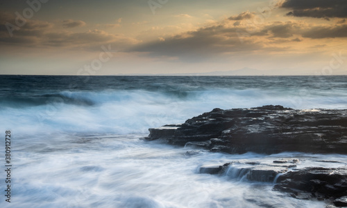 Rocky seashore with wavy ocean and waves crashing on the rocks at sunset. © Michalis Palis