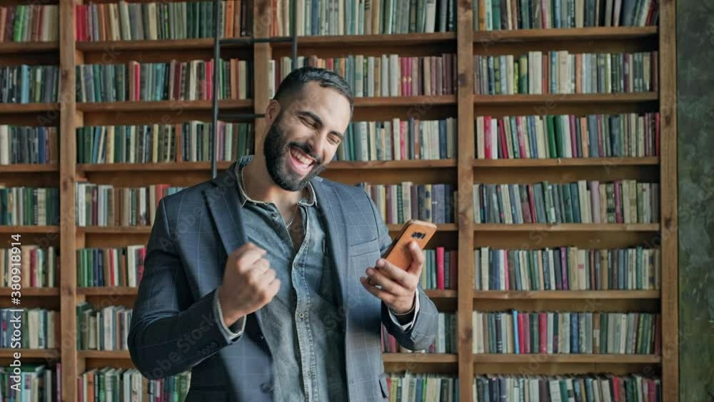 Young Man In The Library With Mobile Phone In His Hands. Something Pleasantly Surprises Him. Young Man In The Library With Mobile Phone In His Hands. Something Pleasantly Surprises Him.