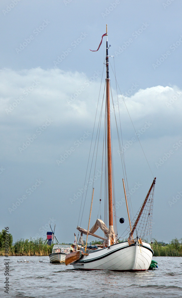 Naklejka premium Wooden sailing boat with lowered sails awaits an approaching rainstorm. In the background a traditional Dutch windmill.
