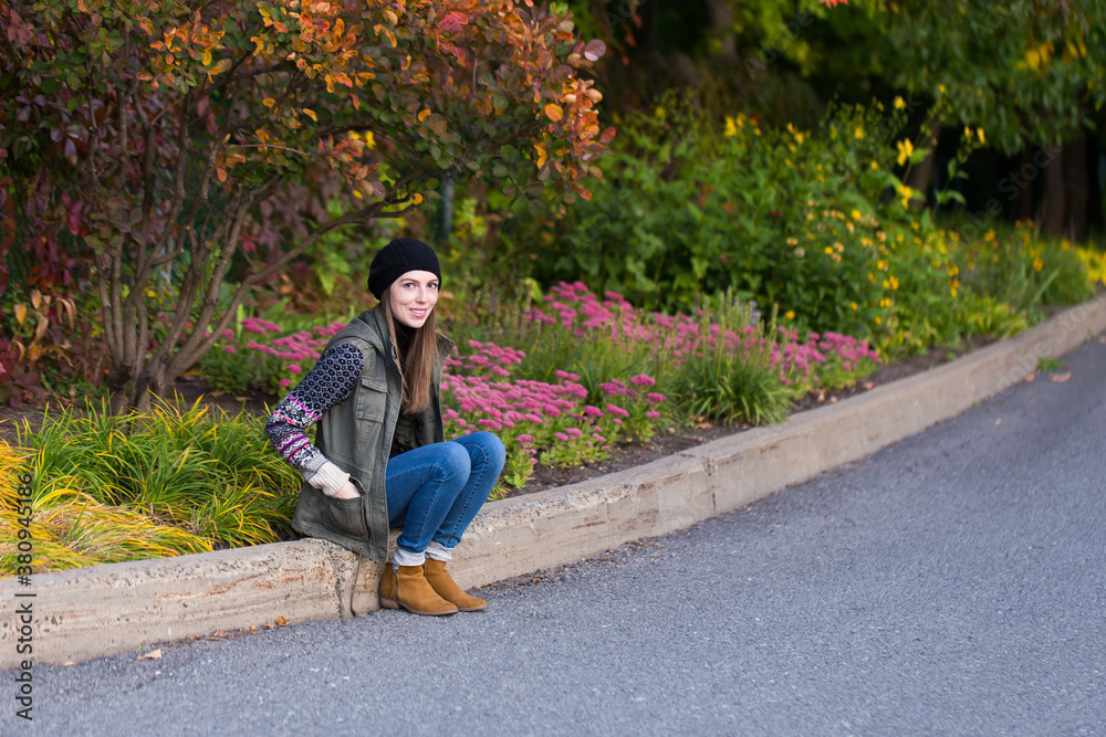 Obraz premium Full length portrait of pretty smiling young woman in casual clothes sitting on a curb during a Fall afternoon, Quebec City, Quebec, Canada