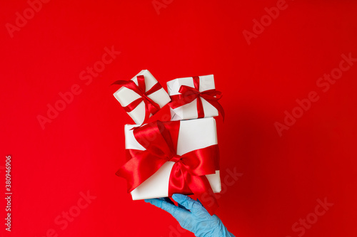Holiday gifts. Pandemic restriction. Greeting congratulation. Hand in protective gloves holding present boxes wrapped taped ruby ribbon isolated on red. Sale discount. Stay safe.