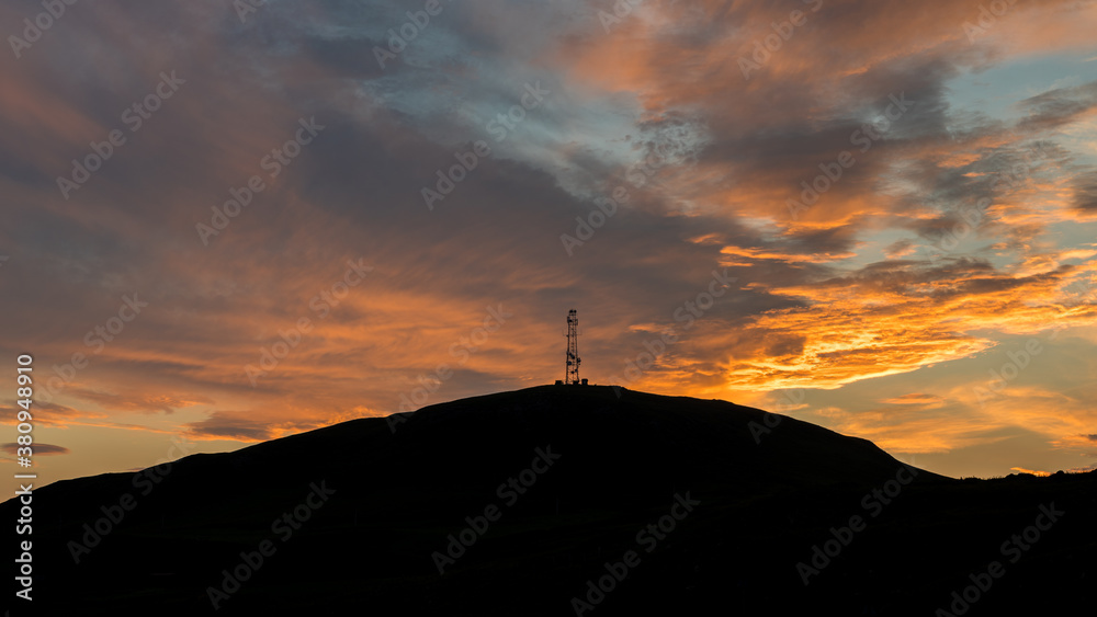 Sunrise behind a hill with a silhouette of transmitter telecommunication antenna on top of hill. Colorful dramatic clouds during sunrise. Sunrise behind a hill with a silhouette of transmitter telecommunication antenna on top of hill. Colorful dramatic clouds during sunrise.