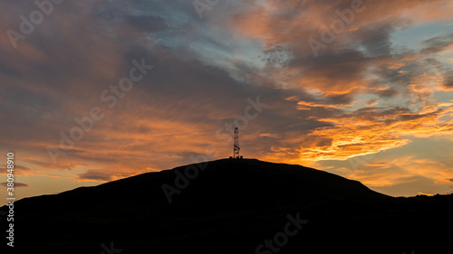 Sunrise behind a hill with a silhouette of transmitter telecommunication antenna on top of hill. Colorful dramatic clouds during sunrise.