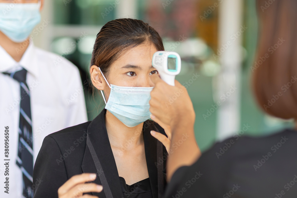 Asian woman teacher using thermometer temperature screening student for fever against the spread of COVID-19 while student standing in queue keep distance before entering into classroom. Asian woman teacher using thermometer temperature screening student for fever against the spread of COVID-19 while student standing in queue keep distance before entering into classroom.