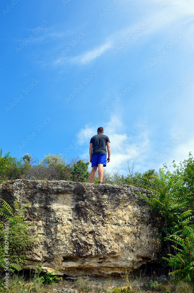 a man stands on the edge of a cliff around green bushes, above the blue sky