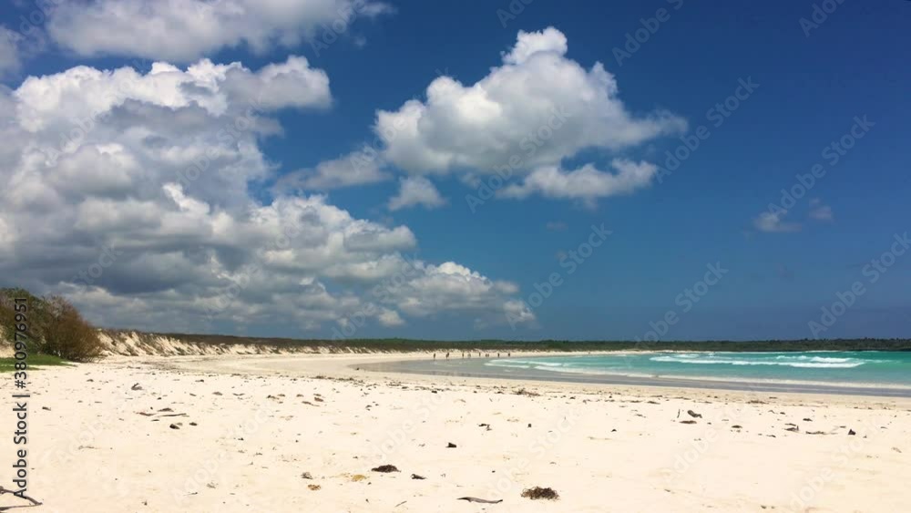A turquoise water with long waves entering the beach with sand creating a reflecting surface for white clouds on a blue sky in Tortuga Bay, Santa Cruz Island, Galapagos.