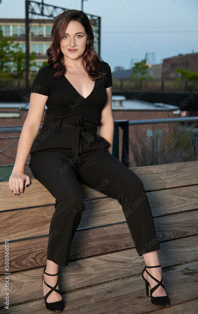 Stunning young woman poses for photo in late summer in elevated urban park - sitting on large wooden swing in black blouse and pants Stunning young woman poses for photo in late summer in elevated urban park - sitting on large wooden swing in black blouse and pants