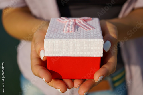 a woman holds a small gift box in beautiful hands with a well-groomed manicure. The girl is outdoors, wearing a light shirt over a black T-shirt 