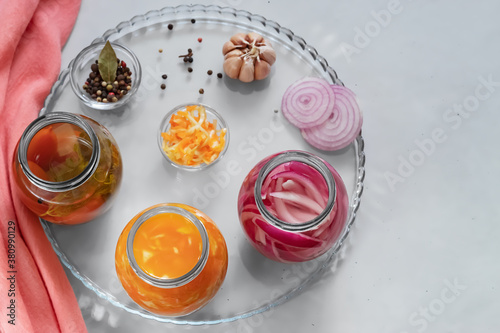 On a glass tray fermented vegetables in jars, next to a bowl with vegetables and various peppers, garlic. A pink napkin. Copy space. Gray background. View from above photo