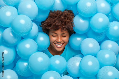 Overjoyed dark skinned woman laughs happily sticks out head through blue balloons happy to celebrate hens party smiles broadly and shows white teeth. Afro American girl prepares for anniversary