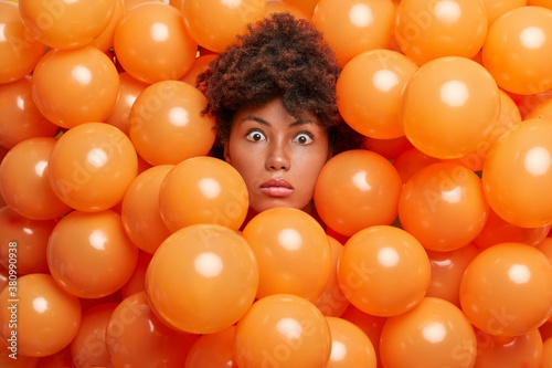 Surprised birthday girl poses over inflated orange party balloons stares bugged eyes expresses great wonder or surprise. Young Afro American woman enjoys anniversary event has shocked expression photo