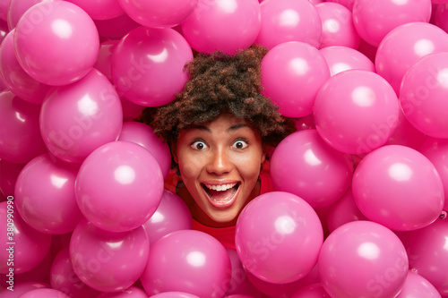 Photo of postive crazy dark skinned woman looks happily at camera gets surprise has fun during festive day surrounded by small inflated rosy balloons. Female model prepares for housewarming party